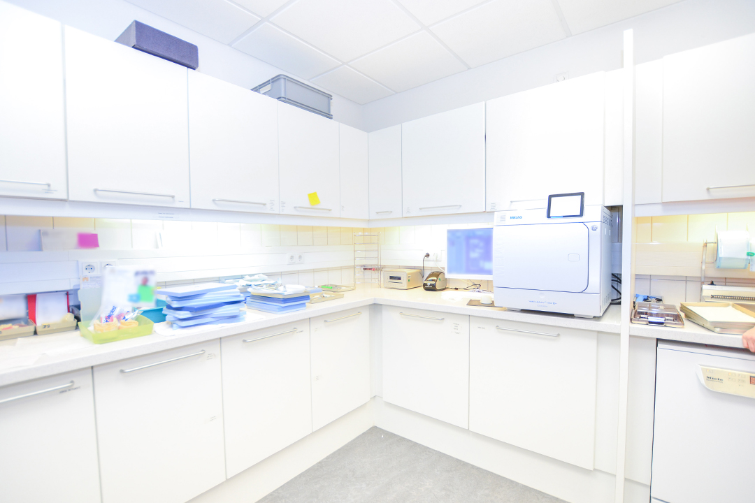Weiße Hängeschränke sowie eine helle Arbeitsplatte sind in einem 90 Grad Winkel angeordnet. Auf der Arbeitsplatte stehen verschiedene Gerätschaften eines Zahnarztes.