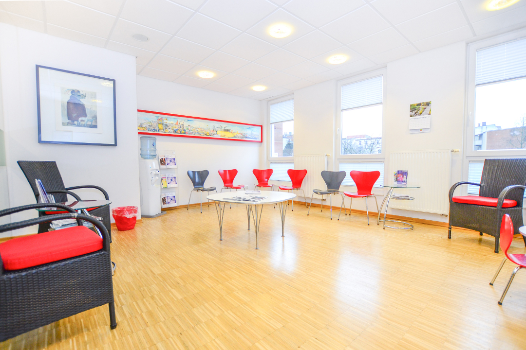 Das Wartezimmer des Flensburger Zahnarztes Dr. Bent Borg. Ein runder Tisch in der Mitte des Raumes ist umgeben von roten und grauen Stühlen, welche in einem abwechselnden Muster aufgestellt sind.